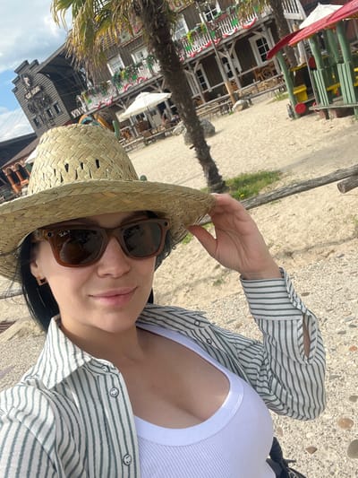 In a selfie taken at a Wild West-themed attraction in Collendoorn, Netherlands, a woman with dark hair wears a straw cowboy hat and sunglasses. She has on a white tank top and a striped button-down shirt, partially unbuttoned to reveal her décolletage. Her right hand adjusts her hat. The background features wooden buildings with balconies decorated with flags, a palm tree, sandy ground, and some playground equipment. The sky is partly cloudy.
