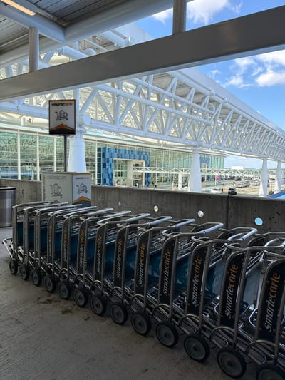 In Charlotte, North Carolina, at Charlotte Douglas International Airport, a line of luggage carts, branded "Smartecarte," is parked on a concrete surface in front of a concrete wall. The carts are silver with black and orange accents. Behind the wall, the modern glass facade of the airport terminal is visible, along with a large, white, truss-like structure overhead that forms a canopy. The sky above is blue with scattered white clouds. Vehicles and people are present in the distance near the airport entrance.