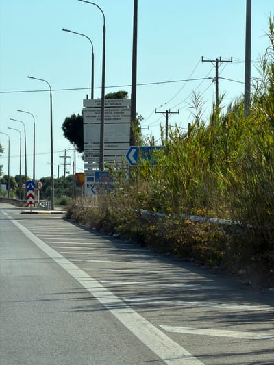 The image shows a road in Areti, Greece, with several streetlights and utility poles visible. A large directional sign with Greek text is situated to the right of the road, and smaller traffic signs are also present. The road itself has white lane markings, and shadows from the trees and streetlights fall across it. The environment appears to be a roadside with overgrown vegetation and a clear blue sky.