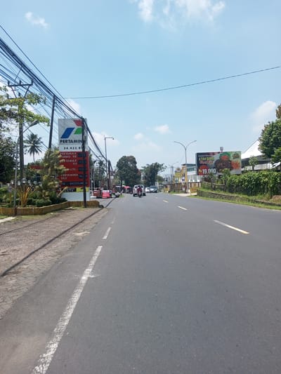 The image shows a street scene, likely in Indonesia given the presence of a Pertamina gas station. The road is asphalt and has lane markings. There is a gas station on the left with a sign displaying gas prices. On the right, there's a billboard advertising Ayam Bakar (grilled chicken) for Rp 19,000. Several vehicles are visible in the distance. Power lines are strung overhead. The sky is clear with some clouds. There is a sidewalk on the left side. Vegetation is present along the road and on the side of the road. There is no indication of this being Nawng Hkar, Myanmar.