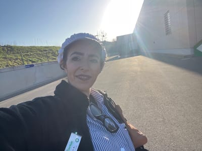 An adult female is captured in a close-up selfie, facing the camera with a slight smile. She is wearing a white, patterned head covering, a dark zippered fleece jacket, and a blue and white striped collared shirt. A pair of black-framed eyeglasses rests on her shirt. An identification badge, displaying the green "Cactus" logo, a green cross symbol, and the partially visible text "Cristina A." and "Caisse," is clipped to her jacket. The outdoor scene is illuminated by bright sunlight, with visible lens flare emanating from the upper right. The background features an asphalt surface. To the left, a concrete retaining wall supports a grassy embankment with sparse vegetation. A blue rectangular sign on the wall displays a white left-pointing arrow and the word "Sortie" (Exit). To the right, a large, modern building with light-colored paneled walls and a vertical window is visible. This scene is situated in Roodt-sur-Syre, Luxembourg, likely at a Cactus supermarket or related commercial facility, as indicated by the identification badge.