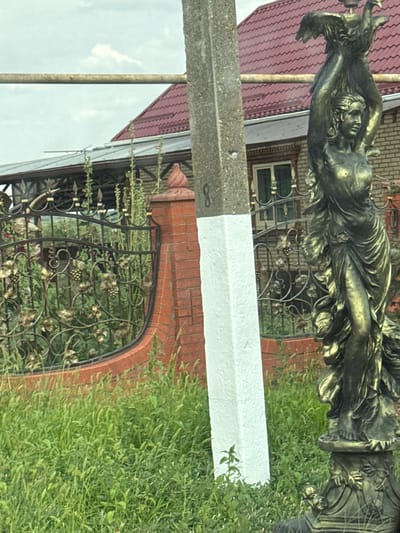 A bronze-colored statue of a woman holding a bird is situated in front of a brick fence and a house with a red tile roof. A concrete pole painted with white stripes stands to the left of the statue. The scene is partially obscured by tall green grass and vegetation. The setting appears to be residential, likely in Kislyakovskaya, Russia, given the architectural style and the presence of a fence common in such areas. The sky is overcast, suggesting a cloudy day. There are no people visible in the image.