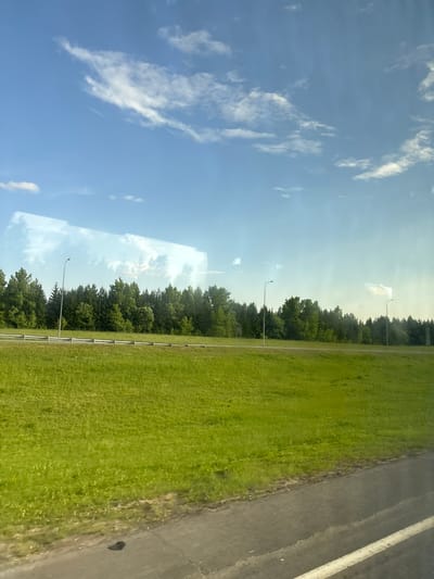 This is a daytime, eye-level, medium shot of a rural roadside scene in Kazan, Russia. The image is taken from a moving vehicle, indicated by the slight blur and reflections on the glass.
The foreground features a section of asphalt road with a white line marking its edge. To the left of the road is a wide expanse of vibrant green grass, likely a roadside verge or field, which appears well-maintained.
Beyond the grassy area, a line of lush green trees forms a dense forest. These trees are varied in height and type, suggesting a natural wooded area. In front of the treeline, a light-colored guardrail runs parallel to the road. Two lampposts, typical of roadside illumination, are visible, one in the left half of the frame and another further to the right.
The sky above is a clear, bright blue, punctuated by scattered, wispy white clouds. The lighting suggests a sunny day, possibly late morning or afternoon. There are subtle, horizontal reflections on the glass, indicating the presence of a window. No people or animals are visible in the frame. The overall impression is one of a peaceful, natural landscape on the outskirts of a city.