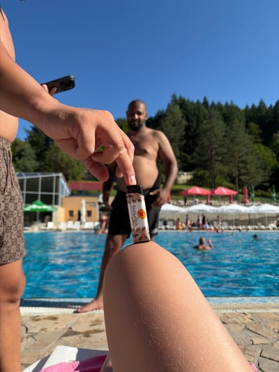 A close-up shot captures a hand holding a small object over a sun-drenched swimming pool at a resort in Chiflik, Bulgaria. The object, a disposable vape pen with "BULGARA" branding and an orange graphic, is the main focus, with a person's thigh prominently in the foreground. In the background, a bald man in swimming trunks stands near the pool, with a dense forest and clear blue sky completing the scene. The pool area is populated with swimmers and lounge chairs, suggesting a leisurely summer day.