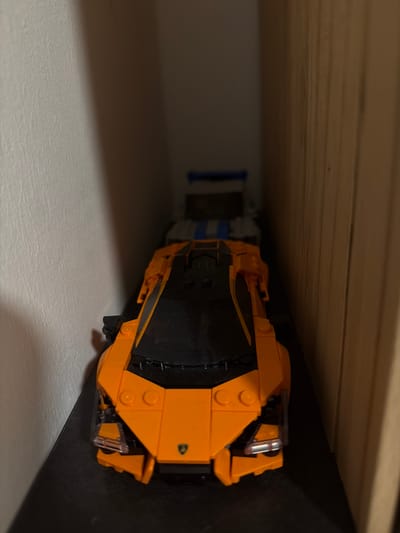 An orange Lego sports car is the main subject in this image, positioned on a dark shelf. The car is detailed with black accents on the hood and body, and appears to be part of a collection as another Lego vehicle, possibly a truck, is visible behind it in the background. The environment suggests an indoor setting, perhaps a room with shelves, with a textured white wall on the left and a wooden surface on the right. The lighting is dim, with a shadow cast across the wall, indicating the image was taken in a low-light condition. The location of Sankt Augustin, Germany is not visually represented in this image.