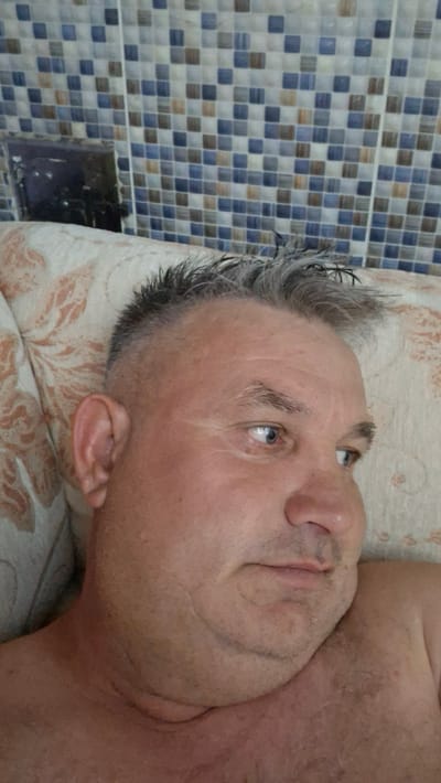 A middle-aged man with short, graying hair and blue eyes is lying on a pillow with a floral pattern, facing to the right. He is shirtless. Behind him, there is a tiled wall with blue and brown square tiles. The overall setting suggests an indoor environment, possibly a bathroom or a bedroom, with no specific indicators of the city of Voinova, Moldova.