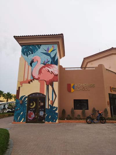 The image depicts a building in Ras Abu Soma, Egypt. The building has a prominent mural of a pink flamingo on a tower-like structure. Next to the mural is a building with the words "Kompass School" and the school logo displayed. A blue electric scooter is parked in front of the school building. The surroundings include paved pathways, shrubbery, and glimpses of palm trees and golf carts in the background. The sky is overcast.