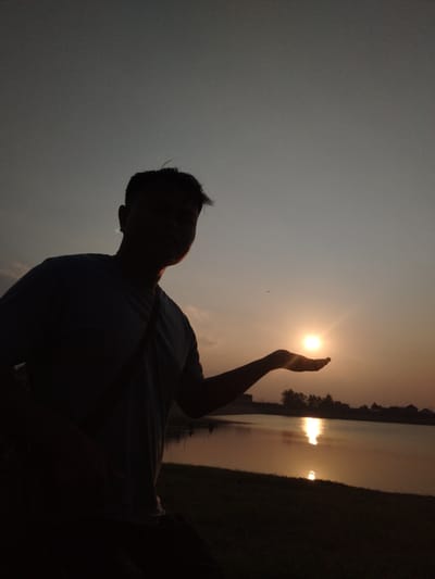 A silhouette of a person extends their hand outwards, catching the setting sun in their palm against a dusky sky. The sun casts a bright reflection on the surface of a calm body of water, likely a lake or river, with the distant silhouette of buildings and trees lining the opposite shore. The ground in the foreground is covered with grass. The image appears to be taken in Blimbing, Indonesia, judging by the context provided.