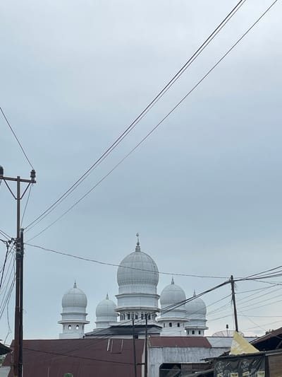 In Jurong Mesjid, Indonesia, under a pale, overcast sky, a white mosque with multiple domes is partially visible behind a rustic corrugated metal roof. The main dome, centrally located, is larger and topped with a crescent moon finial. Several smaller, similarly styled domes flank it. The mosque's architecture suggests a classic Islamic design. In the foreground, a weathered utility pole stands to the left, adorned with insulators and a complex network of dark electrical wires that crisscross the frame, obscuring parts of the sky and the mosque. To the right, another utility pole with wires is visible, alongside a yellow flag and a black banner advertising a product with text and images. The lower portion of the scene shows the reddish-brown corrugated roof of a building or buildings, with some white structures peeking out from behind it. The overall impression is of an urban or semi-urban landscape where modern infrastructure intersects with religious architecture under a muted sky.