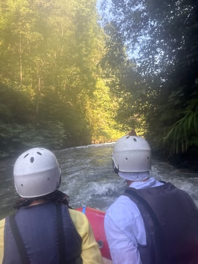 Two individuals, facing away from the camera, are seated in a raft on a flowing river. Both are wearing white helmets and life vests. The person in the foreground, on the left, has dark hair visible beneath their helmet and is wearing a yellow long-sleeved shirt and a blue life vest. The person in the background, on the right, is wearing a white collared shirt and a dark life vest. A red raft is partially visible. The river is turbulent, with white water. The surroundings are densely forested with lush green trees, with some sunlight filtering through the canopy, creating a dappled effect. The scene depicts a rafting activity in a natural, outdoor environment. The location of Rendang, Indonesia is not discernible from the image itself.