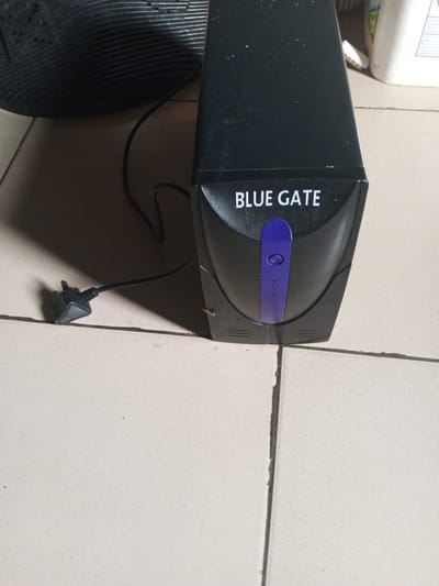 The image shows a black uniterruptible power supply (UPS) unit with "BLUE GATE" printed on it in white text. The UPS has a purple button on the front with three small indicator lights below it. A black power cord with a black plug is attached to the UPS and extends to the left, casting a shadow. The UPS is sitting on a tiled floor, with some dust visible. In the background, a white bottle is partially visible, and a black ribbed object, likely a computer monitor, is on the left side of the frame. The location is identified as Ashata, Nigeria.