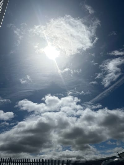 The image captures a bright, sunny day with a partially clouded blue sky. The sun is prominently visible, casting bright rays downwards. Several clouds are scattered across the sky, ranging from wispy cirrus clouds to larger, more defined cumulus clouds. Some of these clouds appear white and fluffy, while others are darker and more shadowed, suggesting varying altitudes or density. In the lower part of the frame, a dark, spiked fence runs horizontally across the scene. Behind the fence, a portion of a dark-colored vehicle and the edge of a roof are visible. The scene appears to be an outdoor location in Tewkesbury, United Kingdom, likely a residential or commercial area, given the presence of the fence and vehicle. There are no people visible in the image.