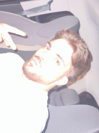 A young adult male with brown hair and a beard sits in a car, holding up a peace sign. The car's interior features gray seats and seatbelts. The location is likely not Rafz, Switzerland, as no geographical or contextual information supports that claim.