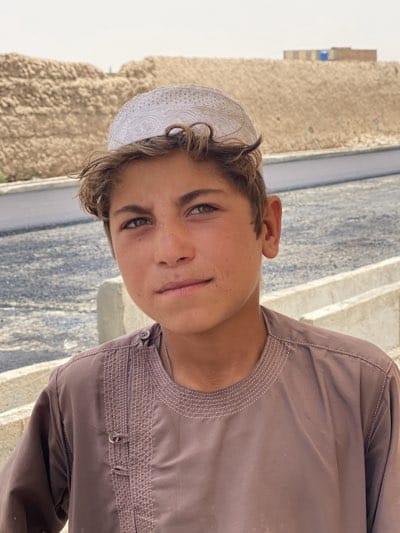 A boy stands outdoors in قندهار, Afghanistan. He is fair-skinned with light brown hair and greenish eyes. He wears a white skullcap on his head and a brown tunic-like shirt with embroidery around the neck and buttons at the shoulder. The background includes a light gray concrete road or walkway, and beyond that, a large mound of tan earth or debris. The scene is well-lit, suggesting a sunny day.