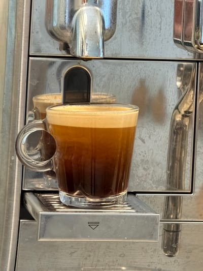 A transparent glass mug of coffee is resting on a metal coffee machine. The coffee has distinct layers, with the top layer a light tan, transitioning to a dark brown. The mug has a clear handle. The coffee machine is made of polished metal and has an arrow symbol on the right side. The background of the image is the metallic surface of the coffee machine. The image was taken indoors in Kfar Saba, Israel.