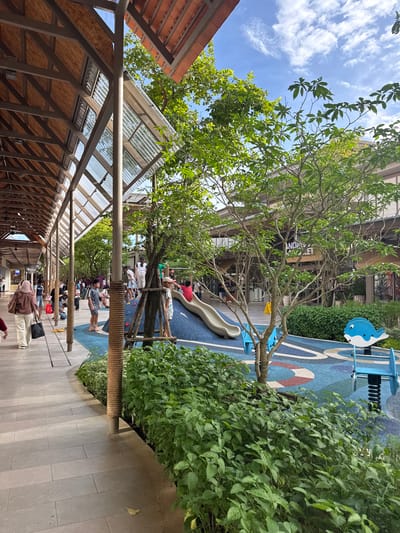 This is a vertical, eye-level, medium-shot photograph taken outdoors in Krabi, Thailand. The scene depicts a modern outdoor shopping area with a covered walkway on the left and a children's play area on the right. The walkway is constructed with a sloped, translucent corrugated roof supported by metal beams, revealing a wooden underside. Paved stone tiles form the walkway.
To the right of the walkway is a vibrant children's play area with a blue, patterned rubberized surface. A white slide is visible, with a few children and adults gathered around it. There is also a blue swing set featuring a stylized blue and white whale design. Lush green foliage surrounds the play area, creating a natural and inviting atmosphere.
Several people are seen walking along the paved area and in the play zone, suggesting a lively, daytime scene. The sky above is blue with scattered white clouds, indicating fair weather. The overall impression is one of a family-friendly, contemporary commercial space integrated with green elements, likely a mall or open-air market. No legible text is visible in the image.
