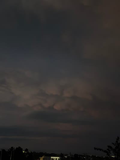 The image depicts a dark, obscured view, possibly taken at night in Novxanı, Azerbaijan. Predominantly, the sky is filled with heavy, dark clouds, some appearing to have a textured, almost turbulent formation. Silhouettes of trees and what appear to be buildings are visible on the right edge of the frame, suggesting the location is at least partially urbanized. A few faint points of light suggest artificial lighting, likely from buildings. The general ambiance is dim and the focus is on the atmospheric conditions.