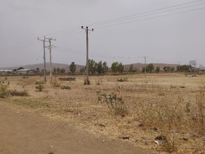 The image shows a dirt road in a rural area, with a water tower visible in the distance. Two electricity poles stand on either side of the road, with a network of power lines strung between them. The ground is dry and sparsely vegetated, with scattered shrubs and small trees. The background is dominated by a hill range, which is visible behind the power lines. The sky is overcast, and the overall impression is of a desolate and somewhat barren landscape. The scene is in the city of Kwadon, Nigeria.