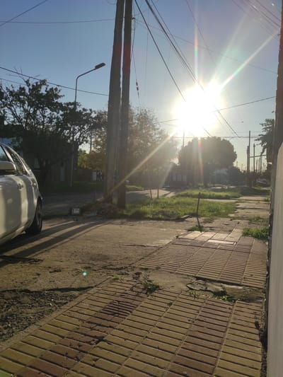 The image depicts a bright, sunny day in Remedios de Escalada, Argentina. The sun is high in the sky, causing significant glare and lens flare. Power lines and wooden utility poles are visible against the blue sky and sunlight. A street lamp is attached to one of the poles.
The foreground shows a brick sidewalk and a paved street. Vegetation grows along the edge of the sidewalk. Part of a white car is visible in the lower part of the frame.
In the distance, trees and buildings are partially obscured by the intense sunlight. A street and more buildings can be discerned further down the road.