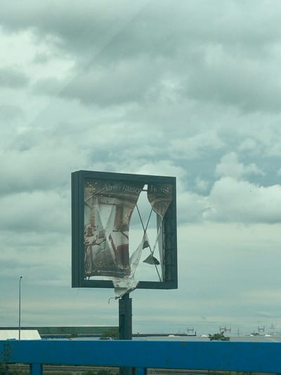The image, oriented correctly, captures a large, severely damaged billboard set against an overcast, cloudy sky. The billboard's advertisement material is torn and shredded, hanging loosely from its frame, suggesting wear from weather or neglect. Fragments of text are visible on the remaining parts of the ad, including "Alpin Reso", "Hotel", "OIANA BRA", "reservation hotel.ro", and "Cel mai mari", implying an advertisement for a resort or hotel. Part of an interior scene, possibly a room or lobby, is faintly discernible on the billboard's surface. To the far right, a sturdy blue structural beam or wall section borders the frame. In the distant background, a flat, hazy landscape stretches towards the horizon, punctuated by several small, indistinct structures or poles. The scene suggests a daytime setting with heavy cloud cover, likely a windy day given the state of the billboard, in an area near Glina, Romania.