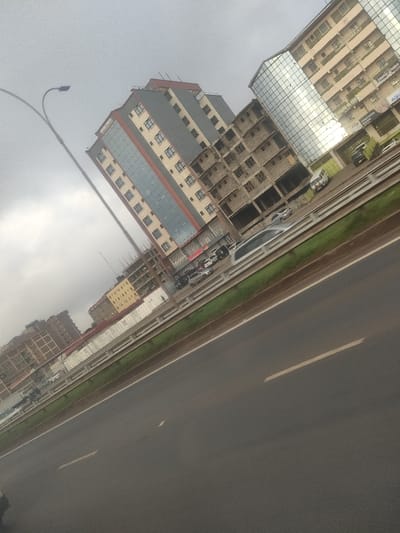 The image depicts an urban landscape in Biashara ward, Kenya, captured at an oblique angle under an overcast sky. In the foreground, a multi-lane asphalt highway is visible with white dashed lane markings. A green strip of grass separates the highway from a raised concrete barrier, behind which a service road or parking area is located.

Multiple vehicles are present: several dark-colored cars (sedans, SUVs) are parked along the service road, and a light-colored van (possibly white or silver) appears blurred, indicating motion. One indistinct pedestrian figure is visible on a sidewalk near the parked vehicles.

The background consists of several multi-story buildings. A prominent modern building with approximately 8-10 floors features a grey facade accented with vertical reflective glass panels and rectangular windows. Adjacent to it, a multi-story structure is under construction, characterized by exposed concrete blocks and visible scaffolding elements. To the right, another multi-story building with a cream-colored facade and numerous windows, some with small balconies, also incorporates reflective glass sections. Ground-floor businesses are visible on this building, including one sign partially legible as "CHEAPEST CAFE." Further down the road, more multi-story buildings, some appearing incomplete or under construction, are visible in the distance. A tall, curved street lamp post with a dark fixture extends from the lower-left into the upper-left of the frame.