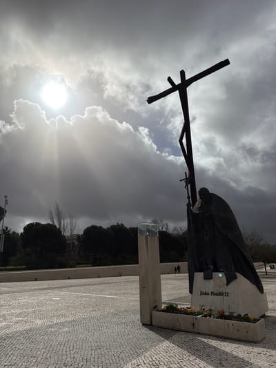 The image depicts the Sanctuary of Fátima in Fátima, Portugal. Central to the foreground is a dark bronze statue of Pope John Paul II, shown kneeling while holding a small crucifix. The inscription "João Paulo II" is visible on the statue's light-colored stone base. Behind and slightly to the left of the Pope's statue stands a taller, dark brown, stylized cross. Both monuments cast prominent shadows onto the expansive, light grey cobblestone plaza. To the left of the Pope's statue, a light concrete pillar with a translucent glass top is present. At the base of the pillar and extending towards the statue are planter boxes containing red, yellow, and purple flowers. In the mid-ground, a low, light-colored concrete wall spans horizontally, with two small, dark human figures walking along it in the distance. A dense line of dark green trees and some bare deciduous trees form the background. The sky is largely covered by substantial grey clouds; however, the sun, positioned in the upper left quadrant, emits intense white light and visible rays through a break in the clouds, creating strong illumination and contrast across the scene.