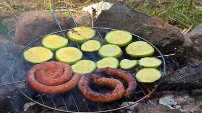 The image shows a close-up, eye-level shot of a round metal grill placed over an outdoor fire pit. On the grill are two coiled sausages and several slices of zucchini. The sausages are a reddish-brown color and appear to be grilled to a crisp. The zucchini slices are green and yellow, with visible seeds, and show some char marks from the grill. Wisps of smoke rise from the grill, indicating that the food is actively cooking. The grill is situated amongst rocks and charred wood, suggesting a rustic outdoor setting. The background consists of dry grass and more rocks. The scene is illuminated by natural light, casting soft shadows. The overall impression is one of an outdoor meal being prepared. The specific location of Dolni Pasarel, Bulgaria is not discernible from the visual information present in the image.