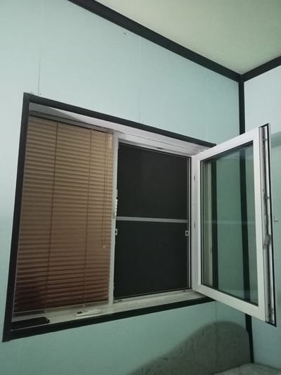 The image shows a partially open window in a room. The window has a white frame and appears to be a double-paned casement window. The left pane is covered by beige horizontal blinds, while the right pane is open and reveals a dark screen behind it. The walls of the room are painted a light blue-green color, and the ceiling is white. A dark frame surrounds the window. On the windowsill, there are two objects: a black rectangular object that could be a phone, and a white remote control. The scene is well-lit, likely by natural light from outside. There are no people visible in the image. The image does not provide any specific visual cues that would allow for the determination of its location in the city of Ryabichev, Russia.