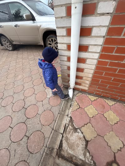 In Gamovo, Russia, a young child, dressed in a dark blue knit hat, a blue quilted jacket with a red inner lining, grey trousers, and black sneakers, stands on a paved surface. The child faces towards the right side of the frame, looking down at the base of a white PVC downspout affixed to a brick wall. The wall is constructed with a pattern of alternating red and light-colored bricks. The ground beneath the child is paved, displaying two distinct patterns: grey concrete with embedded reddish-brown oval elements to the left, and reddish-pink and beige clover-shaped tiles adjacent to the wall on the right. The lower section of the downspout is visibly broken, with damp soil or muddy water pooled around its base. In the upper left background, a dirty silver SUV is parked on the oval-patterned pavement, reflecting an overcast sky and distant trees.
