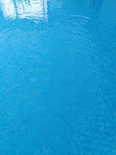 The image depicts the surface of a swimming pool with clear blue water. The bottom of the pool is visible, tiled with a mosaic pattern. Ripples on the water's surface create a shimmering effect, distorting the view of the tiles and reflecting light from above. The scene is devoid of any people or specific objects beyond the pool itself. The setting appears to be an outdoor or indoor pool area, with a hint of overhead structure reflected in the upper portion of the water. The image does not contain any information that would allow for its specific location in Sîngera, Moldova.