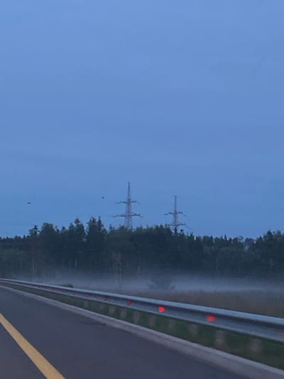The image depicts a scene on a highway in the city of Glinki, Russia, during what appears to be twilight or early morning. The foreground features a paved road with a prominent yellow line marking the lane. A guardrail with reflective red markers runs alongside the road. Beyond the guardrail, there is a stretch of grassy area partially obscured by mist. A dense line of dark green trees forms the background. Two tall electricity pylons are visible in the distance, standing against a pale blue, overcast sky. Two small, red spherical objects are suspended in the sky, positioned at different distances from the viewer.