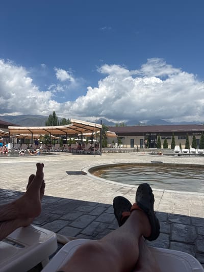 The image depicts an outdoor scene at a spa complex, likely located in Kara-Oy, Kyrgyzstan, given the context. In the foreground, two individuals' legs are visible, resting on lounge chairs. One person's bare feet are crossed, while the other wears black Crocs.

The mid-ground shows a pool area with several people relaxing, some in the water and others on lounge chairs. The pool is surrounded by paved flooring. Beyond the pool, there are shaded areas with awnings, and a building with signage that reads "SPA ZONE" and "SAUNA." Trees and manicured bushes are planted around the complex.

In the background, majestic mountains rise against a bright blue sky filled with scattered white clouds. The overall impression is one of a leisure facility in a scenic mountainous setting.