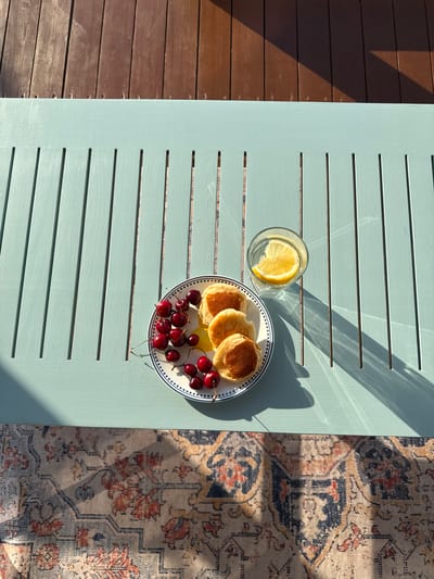 A high-angle shot shows a light blue wooden table with a striped rug partially visible on the right. On the table, a white plate with a blue and white rim holds three small pancakes and a cluster of dark red cherries. To the left of the plate is a clear glass with a slice of lemon in water. The scene is bathed in sunlight, casting long shadows across the table, suggesting it is daytime. The wooden slats of the table and the texture of the rug are visible. To the left of the table, a brown wooden deck is visible, with strong shadows indicating the presence of an overhead structure, possibly a pergola or awning, and the bright sunlight suggests fair weather. The setting appears to be an outdoor patio or deck area. No people are visible in the image, and there is no visible text. The overall impression is one of a peaceful, sunny morning or afternoon setting, possibly in Starokorsunskaya, Russia, given the provided location context.