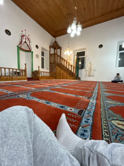 The image depicts the interior of a mosque in Hvostiane, Bulgaria, from a low-angle perspective. In the immediate foreground, the lower legs and feet of an individual, wearing light grey sweatpants and white socks, are visible resting on the floor. The floor is covered by an extensive red carpet patterned with rectangular prayer sections, bordered by blue, white, and intricate geometric designs.

The interior features white walls and a light brown wooden plank ceiling, which includes a central decorative wooden medallion. Illumination is provided by a prominent multi-tiered chandelier with clear glass shades hanging centrally, and a smaller chandelier with three opaque glass shades positioned further back.

On the left wall, a mihrab, an arched prayer niche, is adorned with a red and green crescent moon and star motif at its apex, framed by green borders. To the right of the mihrab, a wooden balustrade runs along the wall. Further into the room, a wooden minbar with a staircase and carved balustrades is positioned.

On the far right side of the room, a male individual is seated cross-legged on the carpet, facing the mihrab. He is wearing a grey hooded sweatshirt and dark trousers, with his hands resting in his lap. Two white robes hang on hooks on the wall near a window, which, along with another window to its left, reveals darkness outside. Circular plaques displaying Arabic calligraphy are affixed to the white walls.