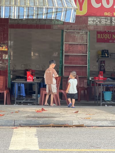 Two young boys are standing on a sidewalk in front of a storefront that appears to be a restaurant or shop. The boy on the left, facing away from the camera, is wearing a brown and white t-shirt and shorts, and appears to be looking at something on a table. He is standing near a red container and some chairs. The boy on the right, facing away from the camera and slightly to the left, is wearing a white t-shirt and black shorts, and is walking away. He has blue flip-flops on his feet. The storefront has red tiled walls and a striped awning overhead. Signs are visible on the wall, one in yellow with red lettering that reads "RƯỢU" which translates to "Alcohol". The sidewalk is paved with concrete tiles and has several fallen leaves scattered on it. The street in front of the sidewalk has a white painted crosswalk. The location is identified as Tân Quang Commune, Vietnam.