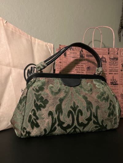 A dark, textured clutch purse with a green and gold damask pattern rests on a dark, flat surface in front of two shopping bags. The purse has a black frame and a black leather top handle with green velvet accents. The shopping bags are behind the purse; the one on the left is a light beige fabric bag with a black circular logo containing the letters "MD". The bag on the right is a pink paper bag with thin pink string handles. The background is a textured, pale gray wall. The scene is indoors and appears to be a still life arrangement. The city of Cape Coral, United States of America is not determinable from the image.