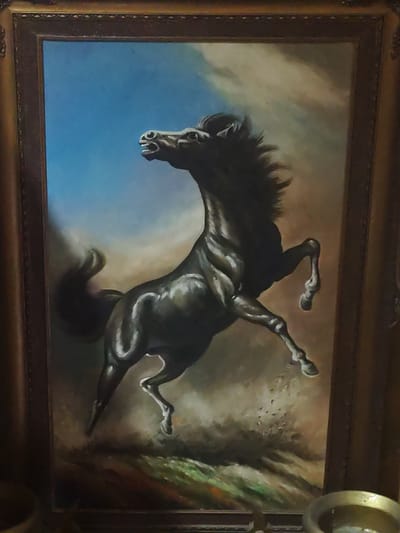 This image, captured in Kokui, Russia, presents a framed vertical oil painting featuring a dark-colored horse rearing. The horse, appearing black or deep brown, is centrally positioned, facing left with its head angled upwards and mouth agape, revealing teeth, indicative of a neigh or cry. Its mane and tail are depicted in a dynamic, windswept manner. The painting's background transitions from a clear blue sky on the upper left to a swirling, cloudy expanse of beige and brown tones on the right, suggesting a dramatic or stormy atmosphere. The lower section of the background shows a textured, earthy terrain, possibly disturbed with dust or debris. The painting is encased in an ornate, gilded wooden frame with sculpted decorative elements. In the foreground of the photograph, at the bottom edge, partial views of two metallic, bowl-like objects are visible, one on the left and a shinier, more reflective one on the right.