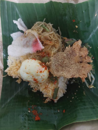 A close-up, overhead shot shows a traditional Indonesian meal served on a banana leaf. The dish consists of rice, a hard-boiled egg with chili flakes, noodles, bean sprouts, and a crispy piece of fried food. The banana leaf is large and green, with visible veins. The meal is arranged in a mound on the leaf, with some chili flakes scattered on the leaf itself. The lighting is natural and soft, highlighting the textures and colors of the food. The background is out of focus, suggesting the photo was taken in a casual dining setting.