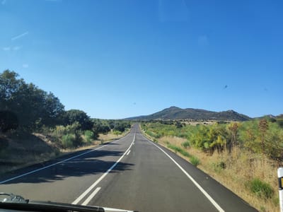 The image shows a view from a vehicle driving on a paved, two-lane road through a rural, mountainous landscape under a clear blue sky. The road is asphalt with white lane markings. On either side of the road are dry grasses, low-lying green bushes, and scattered trees, characteristic of the Mediterranean scrubland. In the distance, a large, rounded mountain range dominates the horizon, its slopes covered with vegetation. The scene appears to be located in or near Santiago de Alcántara, Spain, a region known for its natural beauty and rugged terrain. There are no people or animals visible in the image, and no specific objects or actions are taking place other than the vehicle's movement along the road. The overall impression is one of a peaceful and scenic drive through the Spanish countryside.