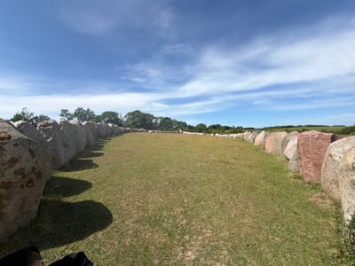 This media file captures a wide-angle view of a megalithic monument, likely a stone circle or burial mound, situated in a grassy field under a bright blue sky with scattered white clouds. The monument consists of a large, curved arrangement of stones, some of which are quite massive and show natural textures and variations in color, including shades of grey, brown, and reddish hues. The stones cast long shadows across the grass, suggesting the photo was taken during the late morning or early afternoon.
The setting appears to be rural or natural, with a line of trees visible in the background beyond the stone structure. The field is covered in dry, yellowish-green grass, indicating a sunny or possibly dry period. The overall atmosphere is serene and historic, evoking a sense of ancient history. While there are no people prominently featured, the lower left corner of the image shows a portion of dark fabric and possibly a person's leg, suggesting someone is present, possibly observing or photographing the site. The location is identified as Lejre, Denmark, a region known for its historical sites. There is no visible text in the image.