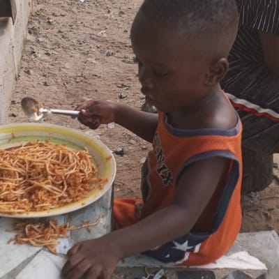 This image depicts a young Black child, approximately a toddler, seated outdoors in Koko, Nigeria. The child wears an orange sleeveless top with "PHOENIX" and a star motif on the front. In the child's right hand, a silver-colored spoon with a dark handle is held above a large, yellow and green-rimmed plate. The plate contains a substantial portion of spaghetti mixed with a reddish sauce and granular components. Several strands of spaghetti have fallen onto a white surface, possibly a piece of paper or cloth, positioned on a low concrete block in front of the child. The child is partially obscured by the plate and their posture suggests active engagement with the food. In the background, to the right, a segment of an individual wearing a dark garment with white stripes and a red accent is visible. The immediate ground surface is composed of packed dirt and small debris, with a light-colored wall or structure visible on the far left.