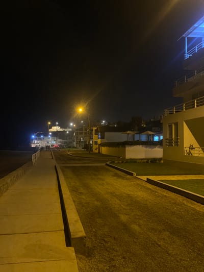 The image shows a street in Punta Hermosa, Peru, at night. It is a dark, nighttime scene, illuminated by streetlights and the glow of the buildings. The street is paved and runs along the beach. A sidewalk on the left side of the image is lit up by streetlights. There are a few people walking in the distance. The street is empty except for a few cars parked on the side. The buildings are modern and well-lit. Some have grassy lawns, while others are surrounded by foliage. The environment is calm and peaceful.