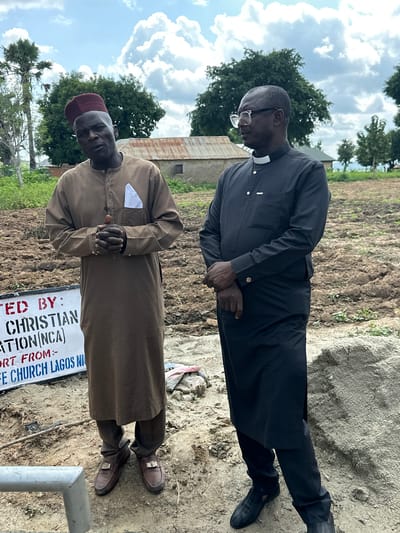 Two men are standing outdoors in Afogo, Nigeria. The man on the left is wearing a tan, knee-length robe with a white pocket square, a red kufi hat, and brown loafers. He is looking at the camera with his hands clasped in front of him. The man on the right is wearing a black cassock with a white clerical collar and black shoes. He is looking off to the side. In the background, there is a dirt field, some trees, and a building with a corrugated metal roof. A sign is partially visible in the foreground, which reads "TED BY: CHRISTIAN ATION(NCA) ORT FROM:- E CHURCH LAGOS NI". The weather appears to be cloudy.