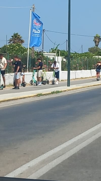 The image depicts a street scene in Ηρακλής, Greece, on a clear, sunny day. The foreground shows a paved road with white lane markings. In the mid-ground, a group of people are standing on a sidewalk next to several electric scooters. A blue flag with the PADI logo is visible in the background, attached to a wooden pole. The surrounding area is lined with a white wall and lush green vegetation, including palm trees. The people appear to be waiting or resting, with some interacting with the scooters. The overall atmosphere suggests a casual outdoor setting, possibly near a tourist area or a rental service.