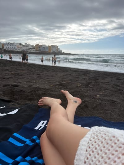 The image captures a beach scene in Puerto de la Cruz, Spain. The foreground features a person's legs and feet resting on a dark blue towel with white and blue stripes. The person is wearing a white, crocheted garment. The feet are bare, and one foot has a small tattoo on the ankle area.
The background reveals a black sand beach with gentle waves rolling onto the shore. In the distance, there is a town with buildings of various colors, including white and yellow, built along the coast. Several people are visible on the beach and in the water, suggesting a relaxed atmosphere. The sky is partly cloudy, with dramatic white clouds interspersed with patches of blue. The lighting suggests it could be late afternoon or early evening. The overall mood appears peaceful and leisurely, indicative of a day at the beach.