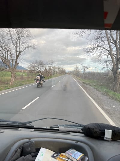 The image shows a view from inside a vehicle, likely a car or truck, driving on a rural road. The road is straight and gray, with a white line marking the lane. A motorcycle is traveling in the same direction ahead of the vehicle. The surroundings consist of fields and bare trees on either side of the road. The sky is cloudy. According to the prompt, the location is near the city of Pevtsite, Bulgaria.