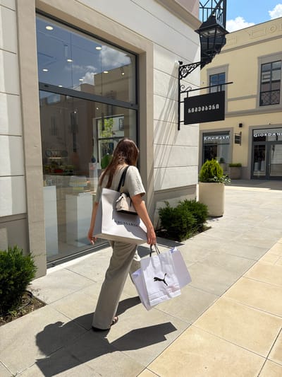 A person walks away from the camera, carrying two shopping bags. One bag, held in the left hand, is a large grey Trussardi bag. In the right hand, the person carries a smaller white Puma shopping bag. The person is wearing a light-colored outfit, including pants and a t-shirt. They are walking on a tiled sidewalk in front of a building with a large glass window. The building has a black sign with white lettering in the Georgian language, and a black wrought-iron lamppost is attached to the building's facade. The environment suggests an outdoor shopping area, possibly an outlet mall. The sign reads "Gamarjveba", indicating the location.