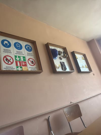 The image shows a wall in what appears to be a building, likely in Dolna Mitropolia, Bulgaria, as indicated by the Bulgarian text on the signs. Three framed displays hang vertically on the wall. The top one features hazard warning signs. The middle display contains a variety of safety equipment, including a hard hat, goggles, gloves, and a respirator. The bottom display is a grid of safety regulation signs, displaying various "mandatory use" and "prohibited" symbols related to hearing protection, eye protection, hard hats, smoking, and hazards for people with implants. To the right of the displays, a metal folding chair is partially visible, mounted on the wall. The wall is a muted orange color.