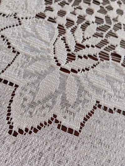 The image is a close-up, high-angle shot of white lace fabric with a floral pattern, laid out on a dark wooden surface. The lace has a detailed, intricate design with openwork sections that reveal the wood grain beneath. The fabric appears textured and slightly raised in certain areas, suggesting a woven or embroidered construction. The lighting is even, highlighting the texture and pattern of the lace. There are no people or active elements present; the scene is static and focused on the textile and its underlying surface. No specific location in Hlynytsi, Ukraine is indicated.