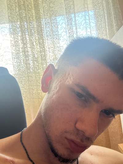 The image is a close-up, low-angle shot of a young man, likely in his late teens or early twenties, with short dark hair and stubble on his chin and upper lip. He has a light complexion and appears to have some acne scarring on his cheek. He is wearing a dark necklace. The man is looking down and to the right, with his face partially in shadow.
In the background, sheer, patterned curtains are visible, suggesting an indoor setting. Sunlight is filtering through the curtains, creating a bright, diffused light. A small portion of a dark chair or sofa is visible on the left side of the frame.
There are no explicit indicators within the image to suggest the location is Gorna Malina, Bulgaria. The scene is domestic in nature, with no identifiable landmarks or signs to pinpoint a specific city or region.